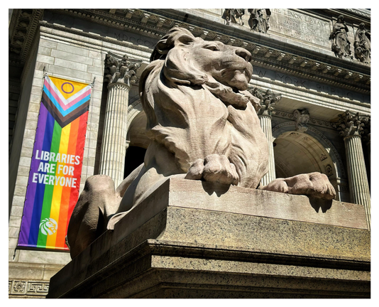 The marble sculpture of Patience the Lion sits regally on his pedestal in front of the ornate facade of the New York Public Library’s 5th Avenue research branch. Behind him and to the left, a long vertical Pride flag hangs from the facade, with the words LIBRARIES ARE FOR EVERYONE printed over the rainbow stripes. Sunlight brings out the highlights and shadows of the sculpture and of the facade’s columns and arches.