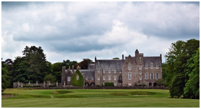 A large stone mansion is set against a backdrop of trees and a cloudy sky. The building features various architectural elements, including towers and large windows, with the 18th green in the foreground.