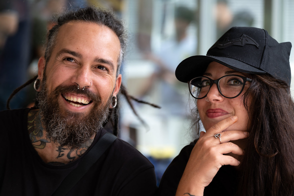Picture of a couple. The man has a beard and mustache, dreadlocks and wears big earrings and a nose piercing. The woman has glasses and waers a cap.