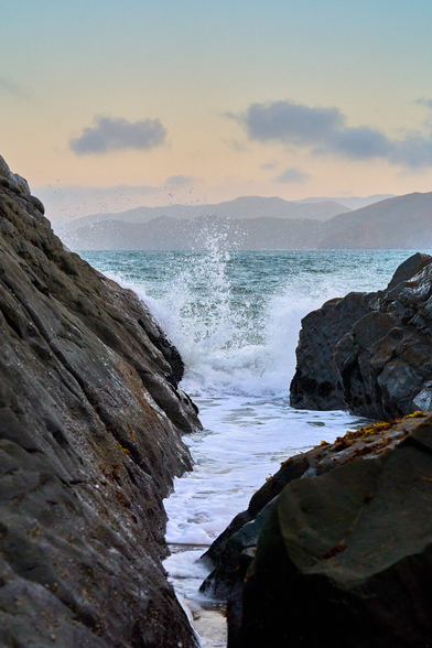 A rocky section of coast looking out to sea. There is a narrow channel with waves crashing at the opening. In the distance are hills.