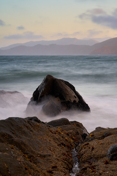 A long exposure of waves breaking on a rock close to shore. The rock is pointed and half covered in mussels. In the foreground is a small drainage channel leading off the rocky shore to the rock. In the far background are the hills of the Marin Headlands. The Pacific Ocean spans the distance.