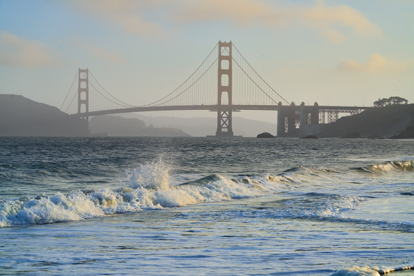 The Golden Gate Bridge backlit and hazy from the sunrise. In the foreground waves are crashing on a beach.