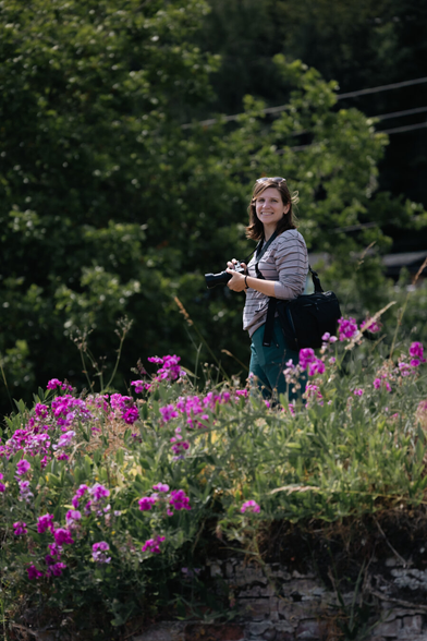a smiling woman standing near flowers, holding a camera
