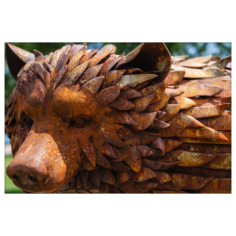 Close-up of a rust-colored metal bear sculpture, with overlapping leaf-like pieces forming the fur and texture of the bear's face and head. The sculpture has a reddish-brown patina with visible weathering, and the expression is stoic with small round eyes and a pronounced snout.