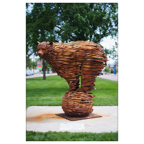 Full-body shot of the same bear sculpture made from curved metal strips, standing on top of a similarly styled metal sphere. The sculpture is placed on a concrete base in a grassy public area, with a tree and urban street in the background. A placard at the base provides information about the art piece.