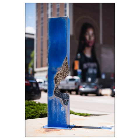 A tall, narrow sculpture resembling a blue steel column with a jagged, organic-shaped cavity running vertically through it. The exposed interior reveals a concrete-like texture. The sculpture is mounted on a concrete base in a downtown area, with a mural of a woman’s face and urban buildings visible in the background.