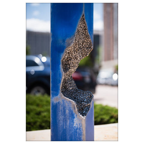 Close-up view of the textured cavity within the blue steel sculpture. The camera focuses on the rough, granular concrete interior and the surrounding worn blue paint, with a blurred urban environment and parked cars in the background.