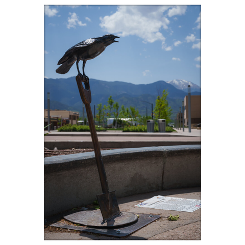 Sculpture of a crow perched on the handle of a metal shovel that is embedded vertically into a metal base. The crow is mid-call with its beak open. The background features mountains in the distance, a mostly clear sky with scattered clouds, and a modern plaza with buildings and young trees.