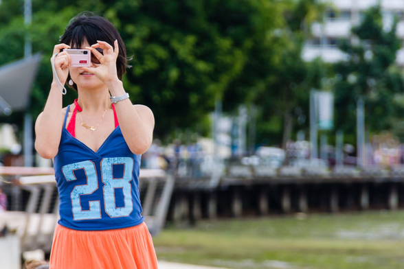 A young woman with a small camera and her hands covering most of her face, looking as if she's about to take a photo. Although she cannot be seen properly, she seems to be young and of Asian appearance. She is dressed in a top with the number 28 on it. The background is out of focus, although it appears there's a wharf with boats behind it and water in front.