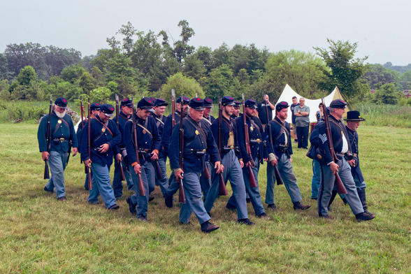 About twenty soldiers march from the rear left towards the front right on closely cut grass with white tent and row of bushes behind them.