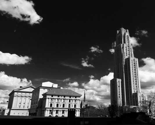 A striking black and white cityscape of Pittsburgh, Pennsylvania, dominated by the Cathedral of Learning, a 42-story Gothic revival skyscraper. The cathedral's intricate stone facade and vertical lines stand in stark contrast against a dramatically dark sky filled with billowing white clouds. In the foreground, several classical-style buildings with columned facades and ornate details can be seen, including what appears to be a government or academic structure. The composition creates a powerful juxtaposition between the soaring modern tower and the more traditional architecture below. The high contrast between the bright clouds and dark sky enhances the image's dramatic and moody atmosphere, emphasizing the city's blend of historical and contemporary elements.
