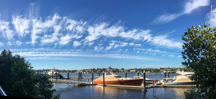 Below is a panoramic shot of the Brisbane river, with boats at moorings, framed by mangroves on the banks. Above is a strange cloudscape. Several thin, linear cloud formations have been blown apart by high winds, creating bands of partially teased out puffballs in lines running almost horizon to horizon. Further in the distance similar linear clouds can be seen, untouched by the wind. The sun is shining and above the clouds the sky is bright blue.
