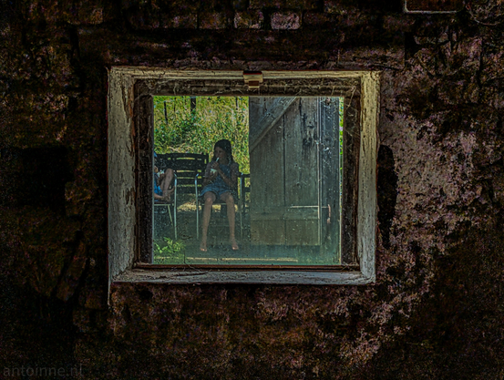 Peering through a filthy window from within a dim, stone barn. The wooden window frame is peeling and worn. The scene outside the window is brighter and reveals two young children sitting on chairs. One girl is sitting towards the centre of the frame, facing slightly to the left, eating an ice cream. The legs of another child are partially visible to the left.