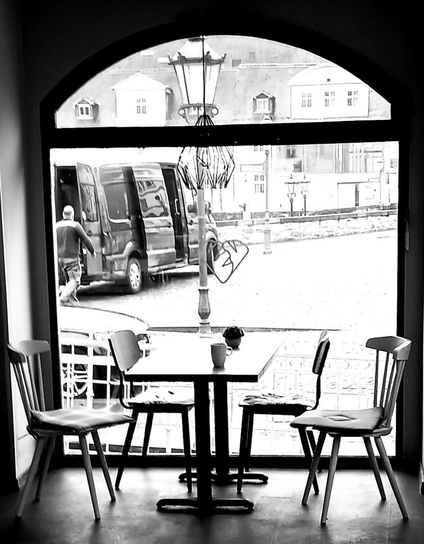 Diese Schwarz-Weiß-Aufnahme zeigt einen Tisch in einem Café mit vier Stühlen; sie alle sind leer und vielleicht haben gerade hier noch Leute gesessen. Auf dem Tisch steht ein Kaffeebecher und etwas Undefinierbares, vielleicht ein Blumengesteck. Die Sitzgruppe befindet sich vor einem großen Glasfenster. Wir erkennen draußen ein Auto, eine Laterne, einen Lieferwagen und eine Person, die uns den Rücken zukehrt, rechts davon einen Platz und dahinter eine Gebäudegruppe.