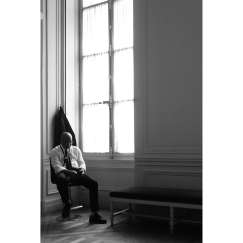 Black and white photo of a museum guard resting on a chair, next to a huge window. The light comes through the old window occupying the third of the picture and illuminates the upper part and the head of the man who gung his jacket behind him. The rest of the picture is darker.