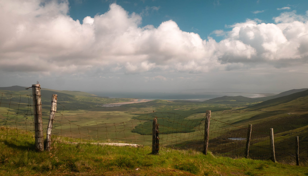 A view of a simple fence on a hill at the top of a mountain pass overlooking the scenery below with a cloudy sky above.