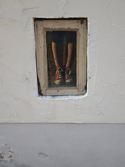 Das Foto zeigt ein kleines, altes Holzfenster, das sich ziemlich niedrig in einer weiß verputzten Wand befindet. Fast gleich darunter fängt schon der graue Kellersockel an. Dass das Fenster schon fast auf Bodenhöhe ist, erkennt man auch an den beiden Beinen in schwarzen Leggings und Stoffsneakern, die sich in der Glasscheibe spiegeln.

The photo shows a small, old wooden window, set quite low in a white plastered wall. The gray basement foundation begins almost directly below it. The fact that the window is almost at floor level is also evident from the reflection of two legs in black leggings and canvas sneakers in the glass pane.