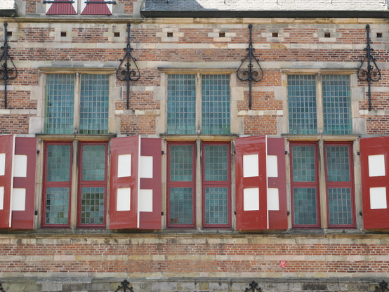 Drei historische Fenster nebeneinander, die unteren Teile haben weinrote Rahmen und ebensolche Fensterläden mit je zwei weißen übereinander angeordneten Quadraten in der Mitte. Drumherum ist Ziegelmauerwerk.