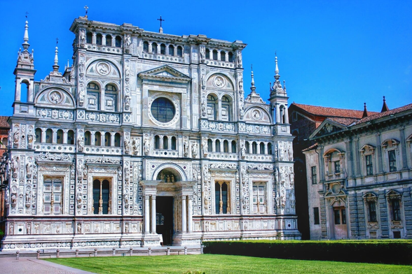 A wide shot of the Certosa di Pavia, a grand Renaissance monastic complex in Italy, under a clear blue sky. The ornate white marble facade of the main church is highly detailed with sculptures, columns, and decorative elements. In the foreground, a manicured green lawn stretches across the bottom of the image, separating the viewer from the impressive structure. To the right, another building with a reddish-brown tiled roof and classic architecture is partially visible.