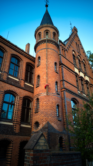 Ein Backsteingebäude im Stil der Neogotik. Neben einem Giebel befindet sich ein Treppenturm mit spitzem Dach. Aus einem Fenster der obersten Etage hängt ein Seil, das unten wie ein Zopf aufgekämmt und mit einer roten Schleife versehen wurde. Das Seil reicht nur etwa bis zur halben Höhe des Turms hinab.