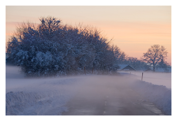 View down a small country lane near the edge of a village. The road has been cleared, there's ~50cm of snow pushed up on either side. A dense, but thin fog bank is drifting across, obscuring the road after a few meters. A group of trees and the first houses are clearly visible further back against the orange/purple sunset sky.