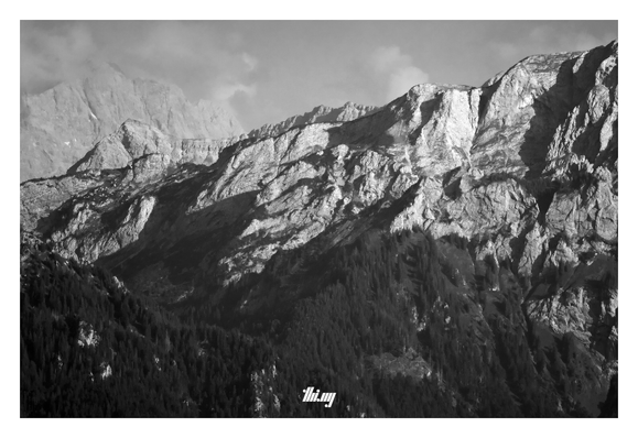 B&W photo of the north-eastern side of Hochplatte with the forested valley in front, and the Zugspitze (Germany's tallest mountain) in the hazy background, its peak surrounded by clouds.