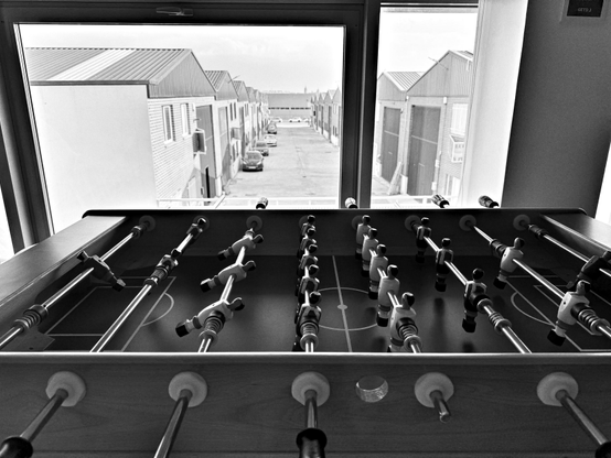 Una mesa de futbolín en blanco y negro se encuentra en primer plano, con sus barras y jugadores de miniatura alineados. A través de una gran ventana detrás de la mesa, se observa una vista de un área industrial con varios edificios de almacenes de un solo piso. Los edificios son sencillos y uniformes.  El enfoque principal está en la mesa de futbolín, con el fondo ligeramente desenfocado pero visible.