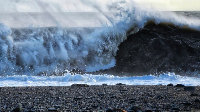 A dramatic close-up of a massive, dark wave crashing and breaking towards a stony beach. The wave's crest is white with foam and spray being blown back by the wind, illuminated by sunlight from the upper right, while the dark body of the wave shows its immense power. The foreground features a pebbled beach stretching across the bottom of the frame.