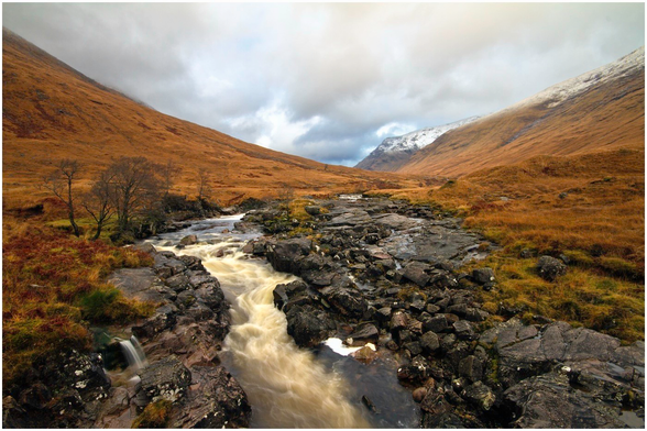 A scenic view of a winding river flowing through rocky terrain, surrounded by rolling hills covered in golden grasses and patches of snow. The sky is overcast with dark clouds.