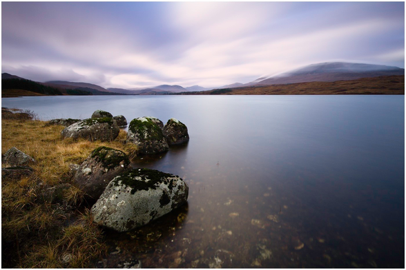 A serene landscape featuring a calm loch bordered by moss-covered rocks and dry grass. In the background, rolling hills and mountains are shrouded in soft clouds, creating a tranquil atmosphere. The sky is partly cloudy, with gentle hues.