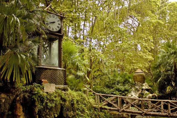 Im Vordergrund eine einfache Holzbrücke, die vom rechten Bildrand zu einer großen Uhr links im Bild führt. Am linken Bildrand eine Palme, im Hintergrund Laubbäume