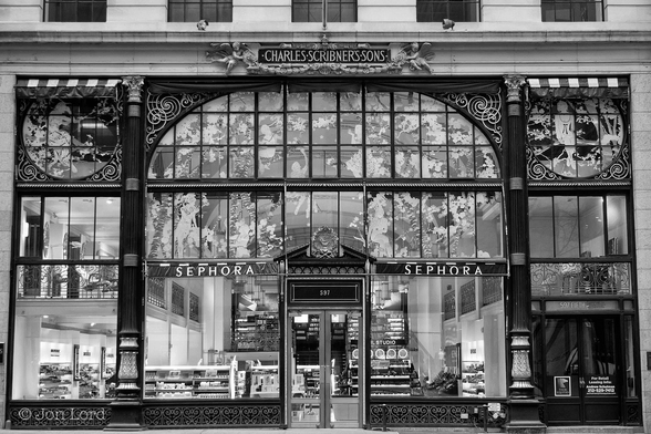 This Is A Black And White Street (But No People) Photo In Landscape Format. Except For A Small (About Ten Per Cent) At The Top: The Entire Image Is Filled With The Ornate Black Wood And Glass Facade Of A Large Shop Front. In The Centre is A Glass Double Door As The Main Entrance And Above Is The Number 597. A Few Metres Either Side Of The Door Are Two Parallel Tall Black Columns Reaching Upwards Three Floors. Towards The Top Of Each Column Is A Decorated Black Fillet Arching Inwards. Filling All This Facade Are Numerous Square And Rectangular Glass Panels. To The Left And Right Of The Columns On The Third Floor Are Four Rounded Fillets, One In Each Corner Making A Circle, Again Filled With Glass Panels. Below Is More Glass Descending To The Ground. Etched White On Much Of The Glass Are Beaux-Arts Style Scenes Of Trees, Shrubs And Women. Above The Facade Is The Name - 'Charles Scribner's Sons'.
The Location: 5th Ave, NYC (2012).