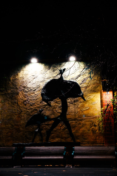 A statue of an odd creature with wing-like arms walking under amber light