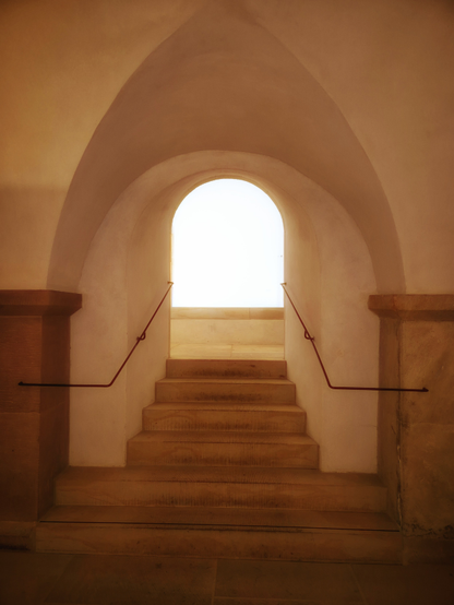 Aus dem Inneren eines Raumes in einem Dom geht der Blick durch einen Rundbogendurchgang in einen hellen Nebenraum. Vor dem Durchgang befindet sich ein Deckengewölbe, das in einem Spitzbogen endet. Eine steinerne Treppe führt zu dem Durchgang, die links und rechts von einem einfachen, dunklen Metallhandlauf aus Rundeisen gesäumt ist.