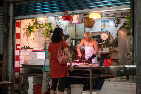 A street-side butcher shop with a butcher at work and customers waiting.