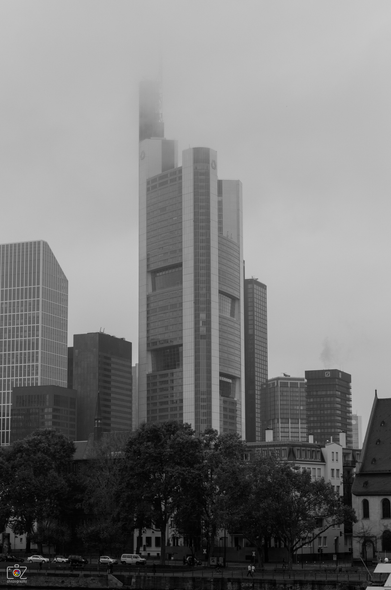 Black and white photo in portrait orientation. In the center is a skyscraper (Commerzbank Tower in Frankfurt am Main), whose upper, characteristic top sinks into the fog. The high-rise is flanked on the left and right by lower office buildings. In the foreground, obscured by trees, residential buildings and a few cars along the (invisible) banks of the river Main.