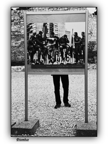 A black-and-white photograph shows an anonymous man, visible from the waist down, standing behind a large display board set up in a public space. The board features a famous image by Chris Niedenthal documenting the strike at the Gdańsk Shipyard in May 1988 — workers sitting on a railing with an industrial landscape in the background. The man is likely observing a different photograph on the reverse side of the display, while for the viewer this juxtaposition becomes a poignant connection between anonymous, everyday presence and the dramatic history of the struggle for freedom.