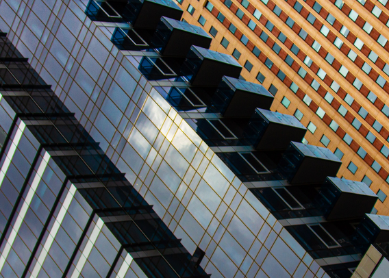 Part of three skyscrapers in New York, photographed at 45 degrees. The left buildings black with the sky and light reflected in them and the last building red brown. and many windows.
