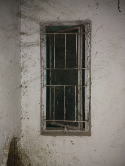 A photo of a small room with an open window surrounded by a white wall covered in dark cobwebs. To the bottom left of the image is a circular decrepit garbage chute.