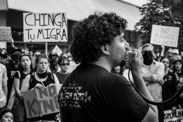 Black and white image of an ICE protest. A man is talking to the crowd through a hand help mic. A sign reading Chinga Tu Migra is in the background. 