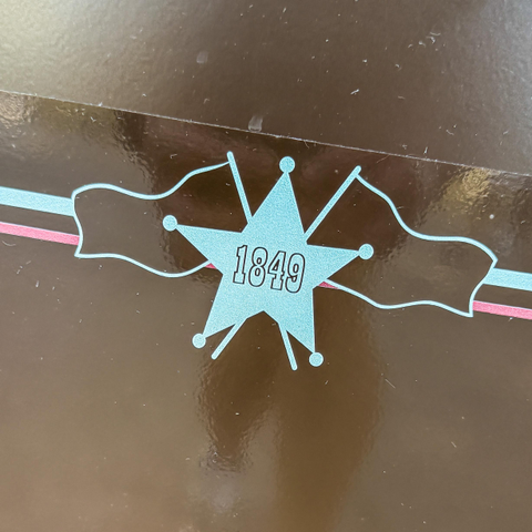 An image featuring a white star with the number "1849" in the center, flanked by two decorative banners, all set against a glossy brown background.
