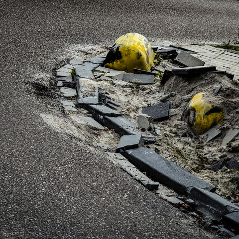 A severely damaged section of road. The scene is dominated by broken asphalt and concrete pieces, scattered amidst loose sand and dirt. The curb of a paved surface is visibly fractured, with individual blocks dislodged and tilted at various angles. There are two distinct, large, roundish objects, yellow and black in color, partially buried in the sand. 