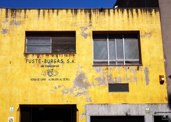 Color photo of the stained and weathered yellow-painted facade of a two story building with two windows on the upper floor. On the wall underneath the window on the left is a painted sign with the words in Catalonian 
“FUSTÉ-BURGAS, S. A.
sala d'especejament
VENDA DE CARN AL MAJOR i AL DETALL” and figures of the heads of a lamb, a cow, and a pig. 