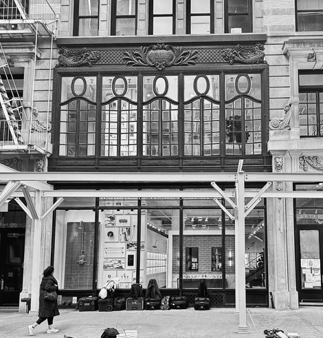  Black and white photo, the lower part shows windows into a bath and tile showroom. In front of that the first layer of scaffolding has gone up as white steel posts on either side and white steel posts across the top. There is a woman walking by looking up and Lots of equipment on the ground from the people who will continue putting up the scaffolding. Right above the scaffolding is a group of five large vertical rectangular windows that are framed in thick black iron and have smaller square black iron grids. Across the top of each window, made of black steel are Loops that look like curly cues that cover the top third of the windows. The building is made of white carved stone on either side and a fire scape can be seen on the left