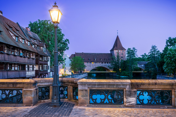 A stone bridge with old street lamp fills the foreground. To the left of frame are timber houses along a blue canal. In the distance is a stone arch and tower resting under a pink sky.