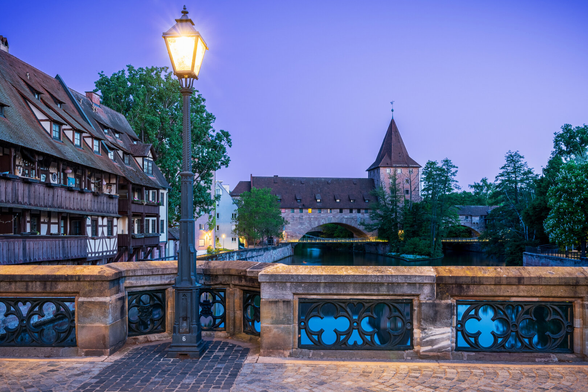 A stone bridge with old street lamp fills the foreground. To the left of frame are timber houses along a blue canal. In the distance is a stone arch and tower resting under a pink sky.