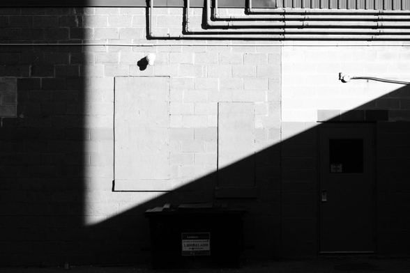 A high contrast black and white image of a exterior wall in the sunlight. Shadows cut the image at a right angle only leaving a triangle shaped area in the center visible. Two windows can be seen boarded up as well as five pipes coming from the right side and then turning up at a sharp angle.