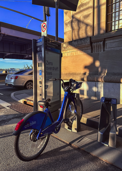 A citi bike parked at a dock on a city street. There’s shadows on a building by the bike. 