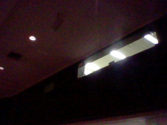 Looking up at a sound booth from a dim theater. The booth is on the 2nd floor and well lit. The bright booth, dark wall, and gray ceiling form harsh geometric shapes.