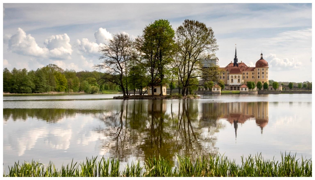 Eine ruhige Seeszene mit dem Barockschloss Moritzburg mit markanten Türmchen, umgeben von Bäumen, die sich im ruhigen Wasser spiegeln. Flauschige Wolken vor Schleierwolken hängen am Himmel. 